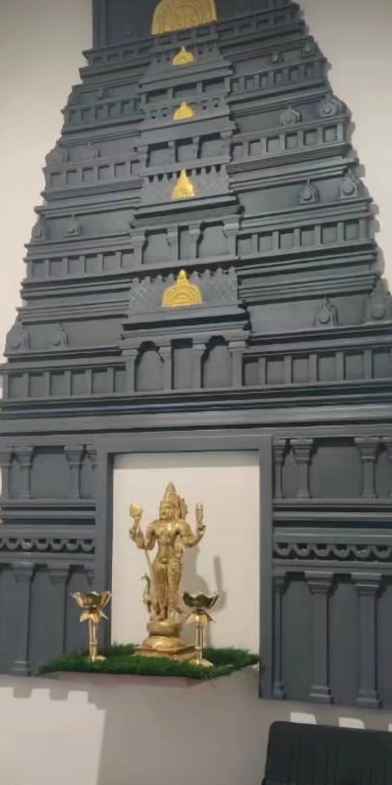 Rawdius Gopuram Customer Installation Dark grey gopuram backdrop framing a brass Murugan idol with oil lamps on artificial turf